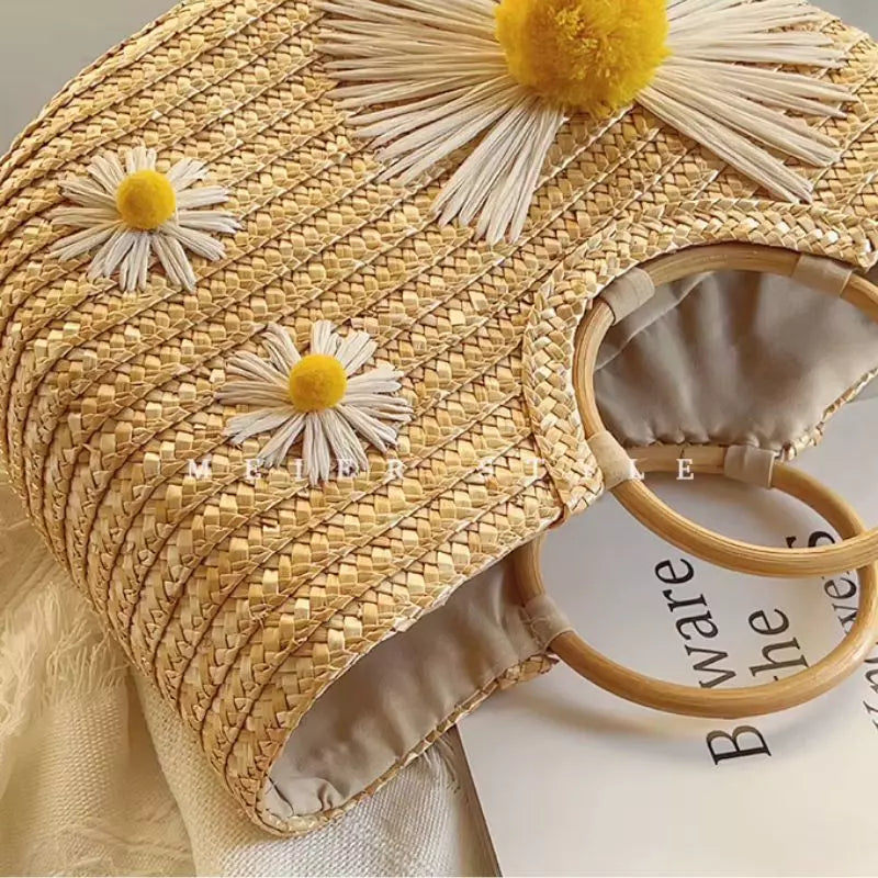 Bolsa de palha deitada de lado na mesa, destacando o relevo das margaridas, as alças de madeira e a abertura interna com tecido.